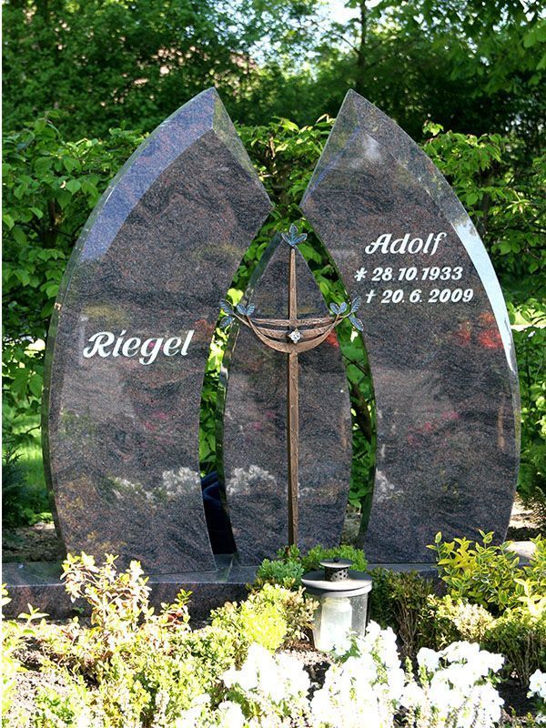Grabstein aus dunklem Stein mit zwei gebogenen, spitz zulaufenden Teilen und einem Kreuz in der Mitte, umgeben von grünen Pflanzen und weißen Blumen